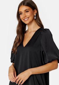 Czarna sukienka z dekoltem w serek i bufiastymi rękawami, wykonana z gładkiego materiału. Charakteryzuje się luźnym krojem i delikatnym blaskiem, noszona z kolczykami w kształcie kropli.