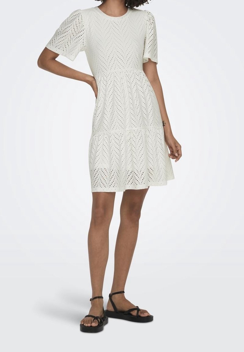 Vit klänning med korta ärmar och rund halsringning, med ett texturerat mönster och en lager på lager, flödande kjol. Bärs med svarta platta sandaler.