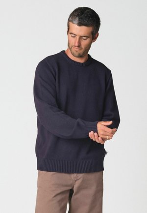 Homme portant un pull bleu marine foncé ajustant la manche avec les deux mains sur un fond clair uni.