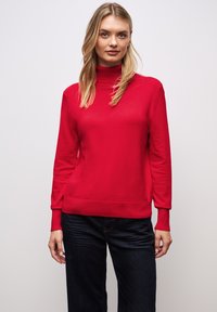 Maglione a collo alto rosso realizzato in tessuto liscio, con maniche lunghe e una vestibilità rilassata. Abbinato a jeans blu scuro.