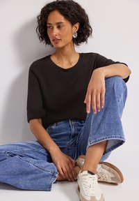 Czarna krótka dzianinowa bluzka w połączeniu z luźnymi niebieskimi dżinsami. Model ma na sobie beżowo-białe sneakersy, siedzi przy neutralnej ścianie.