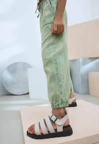 Pantalones de textura verde claro con bolsillos, combinados con sandalias blancas de tira con un cierre en el talón y hebilla, sobre una superficie clara.