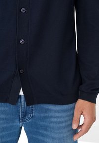 Marineblauwe gebreide cardigan met een gestructureerde afwerking, voorzien van zes ronde knopen en geribde manchetten en zoom. Draagt over een blauwe spijkerbroek.