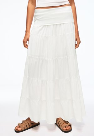 Witte gelaagde maxi-rok gedragen met bruine sandalen met gesp en een witte cropped top, getoond van heupen tot voeten op een staand persoon.