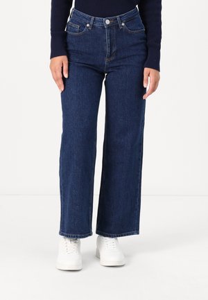 Person wearing dark blue wide-leg jeans, white sneakers, and a navy long-sleeve top, standing against a plain white background.