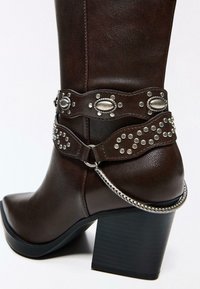 Smeđa kožna čizma do koljena sa šiljastim vrhom, ukrašena dekorativnim smeđim remenom s metalnim detaljima i srebrnim lancem.