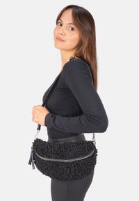 Schwarze Stoffhandtasche mit einer strukturierten, flauschigen Oberfläche, silbernem Reißverschluss und einem abnehmbaren Tragegurt. Verfügt über einen Quastenakzent und eine runde Form.