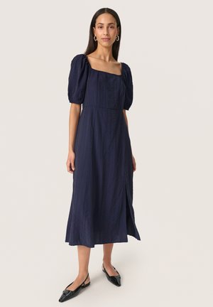 Mujer de pie con vestido midi azul marino con mangas abullonadas, escote cuadrado, abertura lateral, pendientes de aro y zapatos slingback negros con punta.