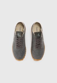 Baskets en suede gris avec un bout arrondi, des lacets foncés et une semelle en caoutchouc marron. Présente une languette de marque et des accents de couture subtils.