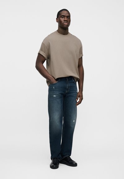 Καφές πουκάμισο με κοντά μανίκια σε συνδυασμό με σκουρό μπλε τζιν με φθαρμένες λεπτομέρειες. Το μοντέλο στέκεται με τα χέρια στις τσέπες, φορώντας μαύρα παπούτσια.