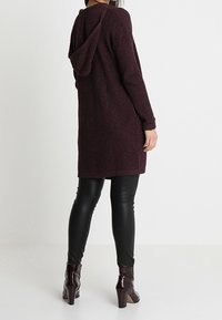 Cardigan en tricot bordeaux avec capuche, longueur moyenne, tissu texturé. Associé à des leggings en cuir noirs et des bottines à talons.