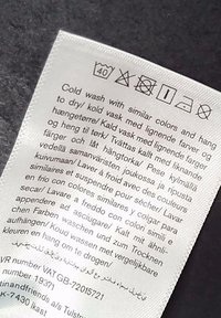 Étiquette d’entretien indiquant les instructions de lavage en plusieurs langues, conseillant un lavage à froid à 40°C avec des couleurs similaires, pas d’eau de Javel, séchage en machine à basse température.