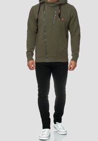 Olivengrüner Zip-Hoodie mit zwei Seitentaschen, strukturiertem Stoff und braunen Lederakzenten; kombiniert mit schwarzen Slim-Fit-Jeans und weißen Sneakers.