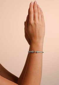 Silverfärgad flätad armband med lås, visad på en hand med fingrar i bönposition. Slät yta, minimalistisk design.