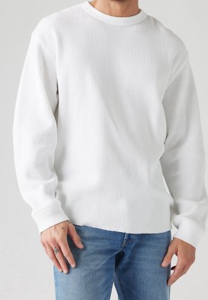 Homme portant un pull blanc à manches longues côtelé et un jean bleu, debout devant un fond uni gris clair.