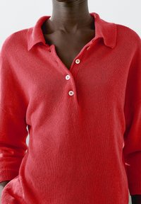 Czerwony sweter z dzianiny z kołnierzykiem, trzema guzikami i długimi rękawami. Tekstura wydaje się miękka i przytulna, o luźnym kroju.