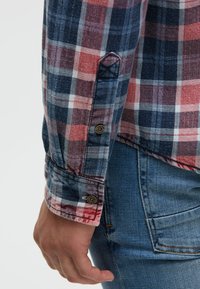 Karohemd in Rot, Marineblau und Weiß; verfügt über lange Ärmel mit Knopfmanschetten und einen strukturierten Stoff. Getragen mit blauen Jeans.