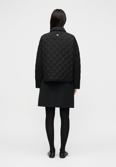 Μαύρο καπιτονέ μπουφάν με μοτίβο σε σχήμα διαμαντιού, με μυτερό γιακά και μικρή λεπτομέρεια λογότυπου στο πίσω μέρος, συνδυασμένο με μαύρη φούστα και καλσόν.