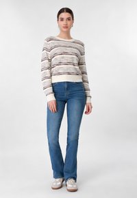 Femme debout portant un pull à manches longues rayé, un jean bootcut bleu, et des baskets blanches et beiges sur un fond blanc uni.