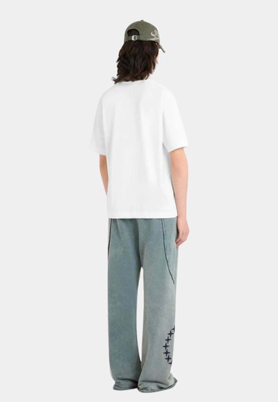 Personne portant un T-shirt blanc surdimensionné, un pantalon de survêtement gris ample avec un motif d'étoile sur la jambe gauche, et une casquette vert olive, tournée de dos.