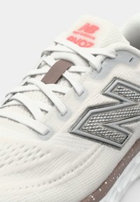 Světle šedá sportovní bota s vrškem z prodyšné síťoviny, texturovanými akcenty, stříbrným logem "N" a růžovým poutkem na patě. Bílá podrážka s hnědými skvrnitými detaily.
