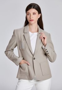 Veste beige à coupe ajustée. Comprend deux poches à l'avant, une fermeture à un bouton et un design décoratif de poche. Texture de tissu lisse.