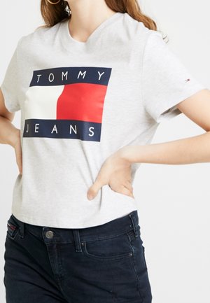 Kobieta ubrana w jasnoszarą koszulkę z dużym logo Tommy Jeans w kolorach czerwonym, białym i niebieskim, w połączeniu z ciemnoniebieskimi dżinsami.
