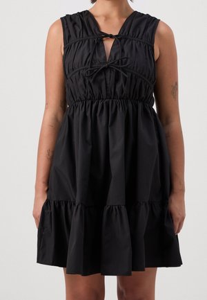 Ärmelloses schwarzes Kleid mit gerafften Stufen und zwei gebundenen Schleifen am Ausschnitt, getragen von einer Person mit sichtbaren Arm-Tattoos.