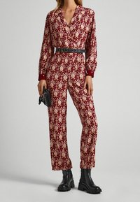 Combinaison à motifs bordeaux et crème, avec des manches longues, un col et une taille élastique. Associée à des bottines noires et un petit sac à main.