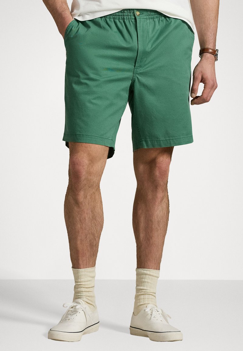 Shorts en coton verts avec une taille élastique, des poches avant et une coupe droite. Associés à des baskets blanches et des chaussettes montantes beiges.