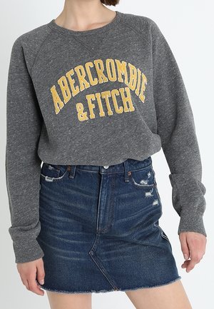 Osoba nosící šedou mikinu Abercrombie & Fitch a modrou džínovou mini sukni s opraným lemem.