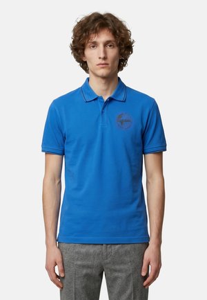 Junger Mann mit lockigem Haar trägt ein königsblaues Poloshirt mit kleinem dunklem Logo auf der linken Brust und graue Hose vor einem einfachen Hintergrund.