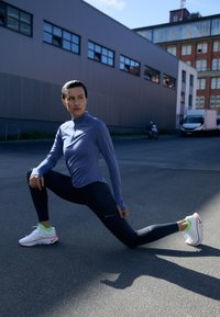 Hosszú ujjú, világoskék sportfelső cipzárral, fekete leggingsszel párosítva. Fehér sportcipők neon zöld és piros részletekkel. Aszfalt háttér.
