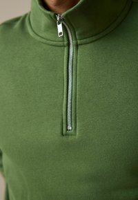 Green pullover made of soft fabric, featuring a high collar and a half-zip front with a silver zipper and textured stitching detail.