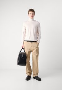 Col roulé rose clair, pantalon beige, ceinture noire et sac en cuir noir. Le mannequin se tient devant un fond neutre et porte des chaussures noires.