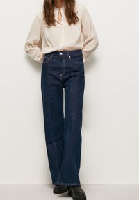 Jeans in denim blu scuro a gamba larga con vita alta, dotati di tasche frontali e dettagli di cucitura sottili, abbinati a una blusa leggera e testurizzata.