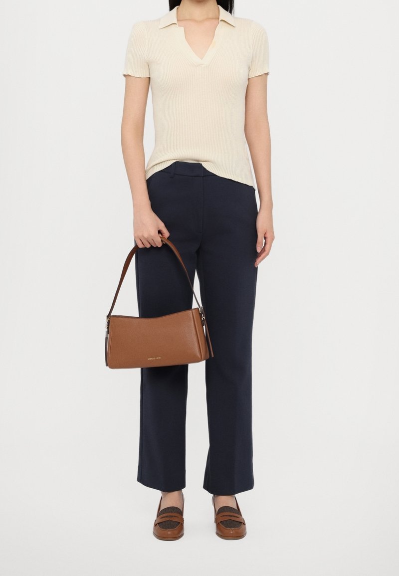 Femme debout, portant un polo à manches courtes crème, un pantalon bleu marine, des mocassins marron et tenant un sac à bandoulière marron devant un fond uni.