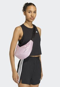 Borsa a tracolla rosa con una texture liscia, forma curvata e tracolla nera, indossata sopra una canotta nera senza maniche e pantaloni corti neri con strisce bianche.