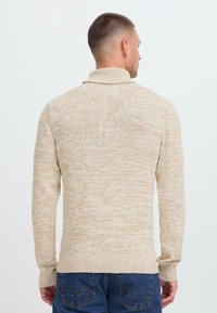 Hombre visto desde atrás, con un suéter de punto beige de cuello alto y vaqueros azules, contra un fondo claro y liso.