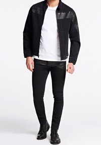 Mustaharma denim -takki kiiltävillä olkakeldoilla, yhdistetty valkoiseen t-paitaan ja mustiin farkkuihin. Mustat nauhakenkäsaappaat viimeistelevät asun.