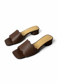 Ζευγάρι καφέ δερμάτινων mule με ανοιχτή μύτη, με κυματιστό περίγραμμα και χαμηλά τακούνια τύπου block, εσωτερικές σόλες σε μπεζ χρώμα, σε λευκό φόντο.