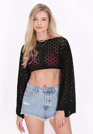 Giovane donna con lunghi capelli biondi che indossa un top corto in pizzo nero sopra un bralette rosa e shorts di jeans azzurri a vita alta.