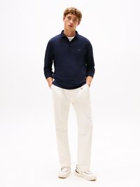 Polo de manga longa azul-marinho com colarinho, apresentando um pequeno logotipo. Combinado com calças brancas e ténis brancos, em fundo simples.