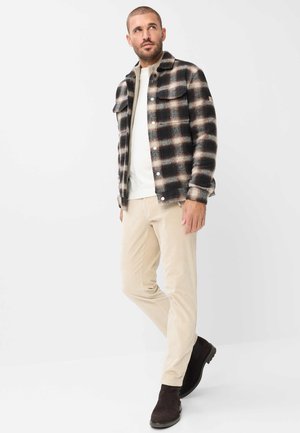 Mand iført en sort og beige ternet jakke, hvid skjorte, beige bukser og sorte støvler, stående med det ene ben krydset foran det andet.