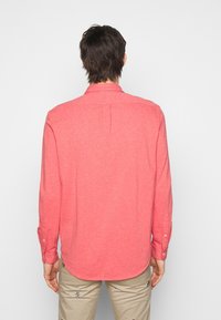 Camicia rossa a maniche lunghe con colletto a bottoni, tessuto strutturato, pence sul retro e polsini con bottoni, abbinata a pantaloni di colore chiaro.