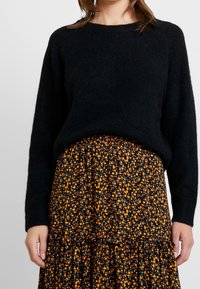 Een vrouw draagt een zwart, lang mouwtrui die is ingestopt in een zwarte rok met een klein oranje en witte bloemenpatroon.