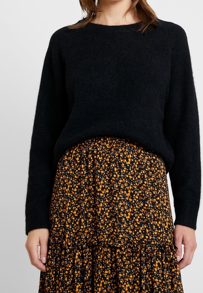 Een vrouw draagt een zwart, lang mouwtrui die is ingestopt in een zwarte rok met een klein oranje en witte bloemenpatroon.