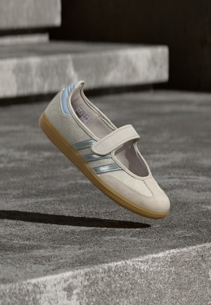 Beige tøysko med elastisk stropp, teksturert overdel, sølv- og blå detaljer, og en såle av gummigummi, vist mot en grå bakgrunn.