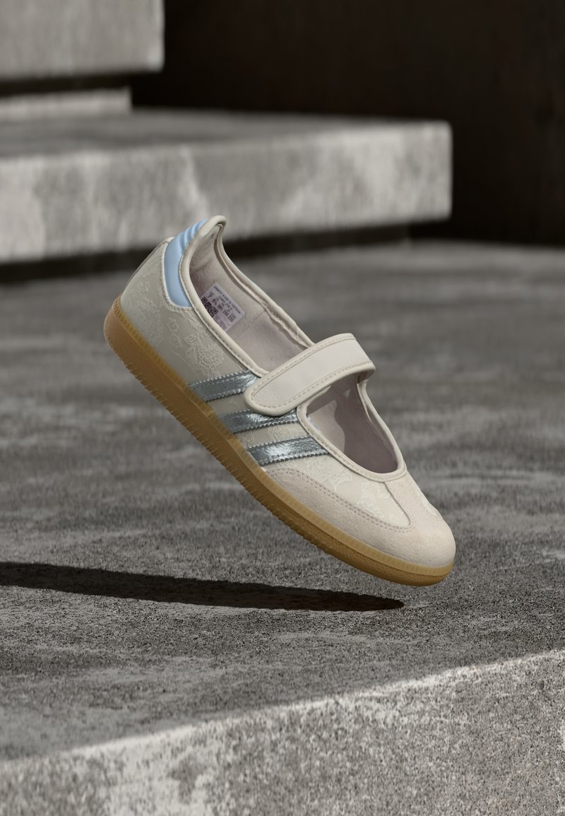 Chaussure en tissu beige avec sangle élastique, dessus texturé, accents argentés et bleus, et semelle en caoutchouc gomme, présentée sur un fond gris.