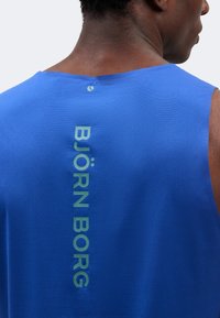 Άνδρας που φοράει μπλε αμάνικη μπλούζα με κάθετο κείμενο "BJÖRN BORG" στην πλάτη, εμφανίζεται από τον λαιμό μέχρι το μέσο της πλάτης.